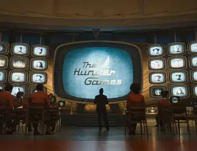 A large screen displays "The Hunger Games" emblem surrounded by numerous smaller screens with portraits. Silhouetted viewers face the screens in a dimly lit room with wooden furnishings. A standing figure is centrally framed, emphasizing suspense and anticipation in the setting.