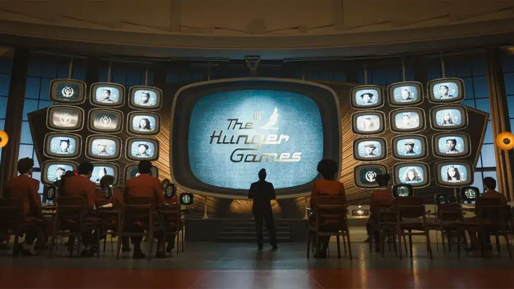A large screen displays "The Hunger Games" emblem surrounded by numerous smaller screens with portraits. Silhouetted viewers face the screens in a dimly lit room with wooden furnishings. A standing figure is centrally framed, emphasizing suspense and anticipation in the setting.