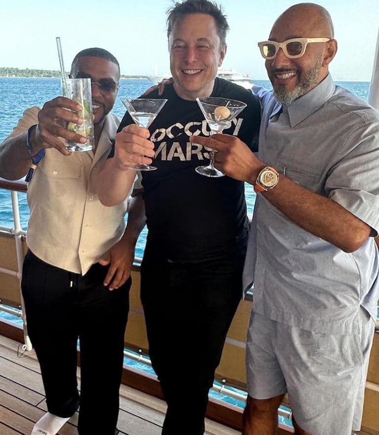 Timbaland, Elon Musk, Swizz Beatz, Three men are standing together smiling. The man on the left is wearing a light-colored shirt. The middle man is wearing a black shirt with "OCCUPY MARS" printed on it. The man on the right is wearing a light-colored shirt and white-framed sunglasses. All are in a sunny outdoor setting with a body of water in the background., Three men stand on a boat deck, smiling and holding drinks. The man in the center wears a "Occupy Mars" T-shirt, the man on the left holds a tall glass with a green drink, and the man on the right, wearing sunglasses and a matching set of a light blue shirt and shorts, holds a martini.