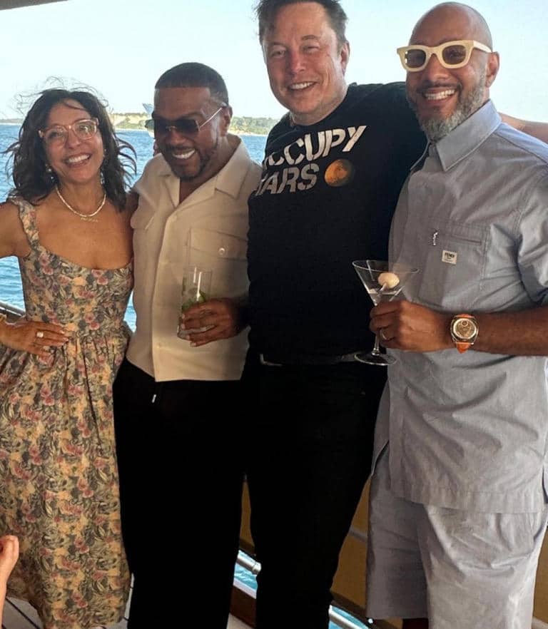 Four people are standing arm-in-arm on a boat with water in the background. The first person is a woman in a floral dress and glasses, the second is a man in a white shirt with a drink, the third is a man in a black "Occupy Mars" t-shirt, and the fourth is a man in a light blue outfit and sunglasses.

.