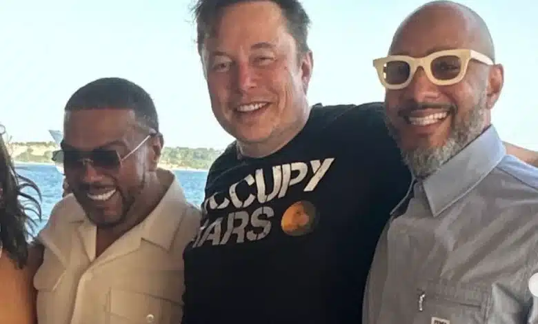 Timbaland, Elon Musk, Swizz Beatz, Three men are standing together smiling. The man on the left is wearing a light-colored shirt. The middle man is wearing a black shirt with "OCCUPY MARS" printed on it. The man on the right is wearing a light-colored shirt and white-framed sunglasses. All are in a sunny outdoor setting with a body of water in the background.