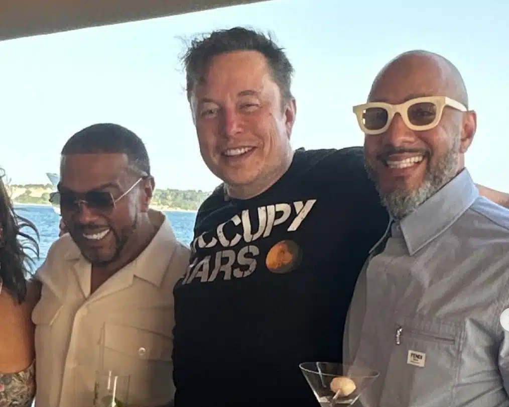 Timbaland, Elon Musk, Swizz Beatz, Three men are standing together smiling. The man on the left is wearing a light-colored shirt. The middle man is wearing a black shirt with "OCCUPY MARS" printed on it. The man on the right is wearing a light-colored shirt and white-framed sunglasses. All are in a sunny outdoor setting with a body of water in the background.
