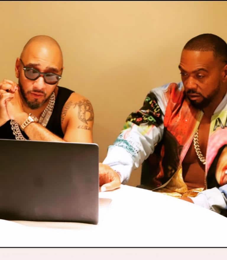 Timbaland, Swizz Beatz, Two men sit at a table focused on a laptop screen. The man on the left wears sunglasses, a sleeveless shirt, and a silver chain necklace. The man on the right wears a colorful jacket and another chain necklace. Both have serious expressions as they look at the laptop.