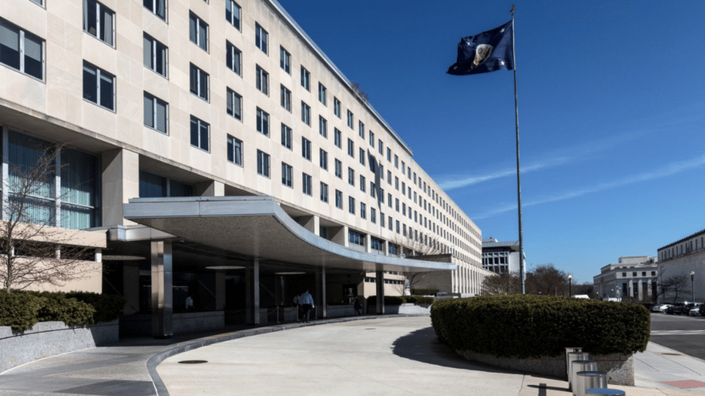 United States State Department, A multi-story government building with a curved, covered driveway in the foreground. The building has many windows and an American flag flies on a tall flagpole to the right. The sky is clear and blue, and there's minimal pedestrian activity near the entrance. Bushes line the sidewalk.