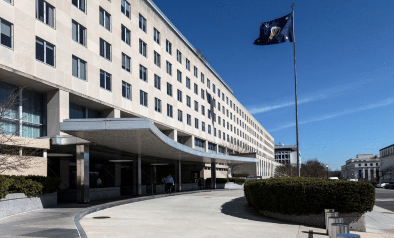 United States State Department, A multi-story government building with a curved, covered driveway in the foreground. The building has many windows and an American flag flies on a tall flagpole to the right. The sky is clear and blue, and there's minimal pedestrian activity near the entrance. Bushes line the sidewalk.