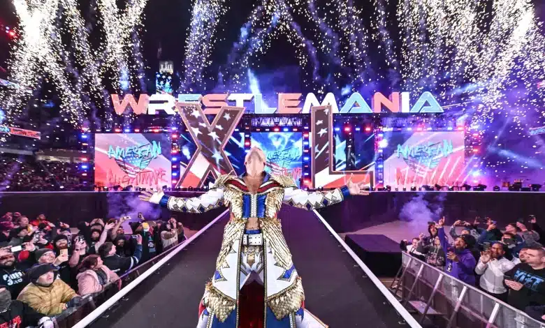 A wrestler stands triumphant on a stage at WrestleMania with arms outstretched, amid a dazzling fireworks display. The stage is illuminated with vibrant lights and the audience surrounds the walkway, cheering. The WrestleMania logo prominently arches over the stage in the background.