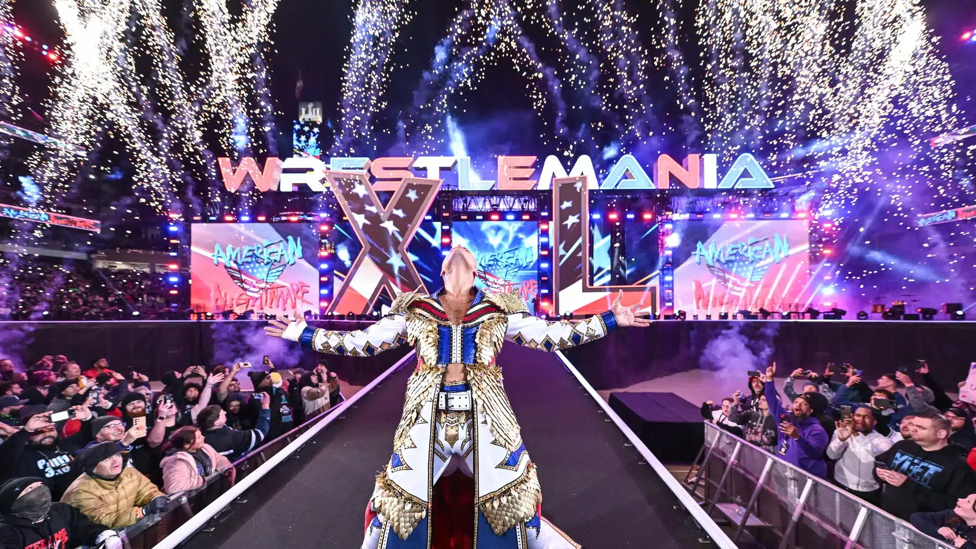 A wrestler stands triumphant on a stage at WrestleMania with arms outstretched, amid a dazzling fireworks display. The stage is illuminated with vibrant lights and the audience surrounds the walkway, cheering. The WrestleMania logo prominently arches over the stage in the background.