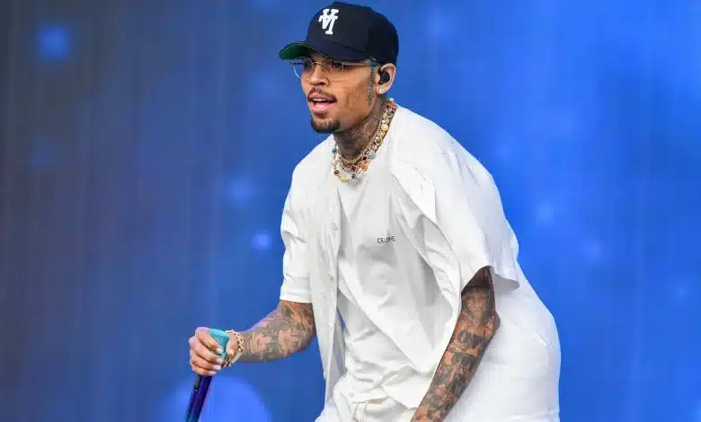 Chris Brown, A man with tattoos, wearing a navy blue baseball cap with "V" on it, glasses, a white short-sleeved shirt, and a white undershirt, performs on stage against a blue background, holding a purple microphone. He has multiple necklaces and bracelets.
