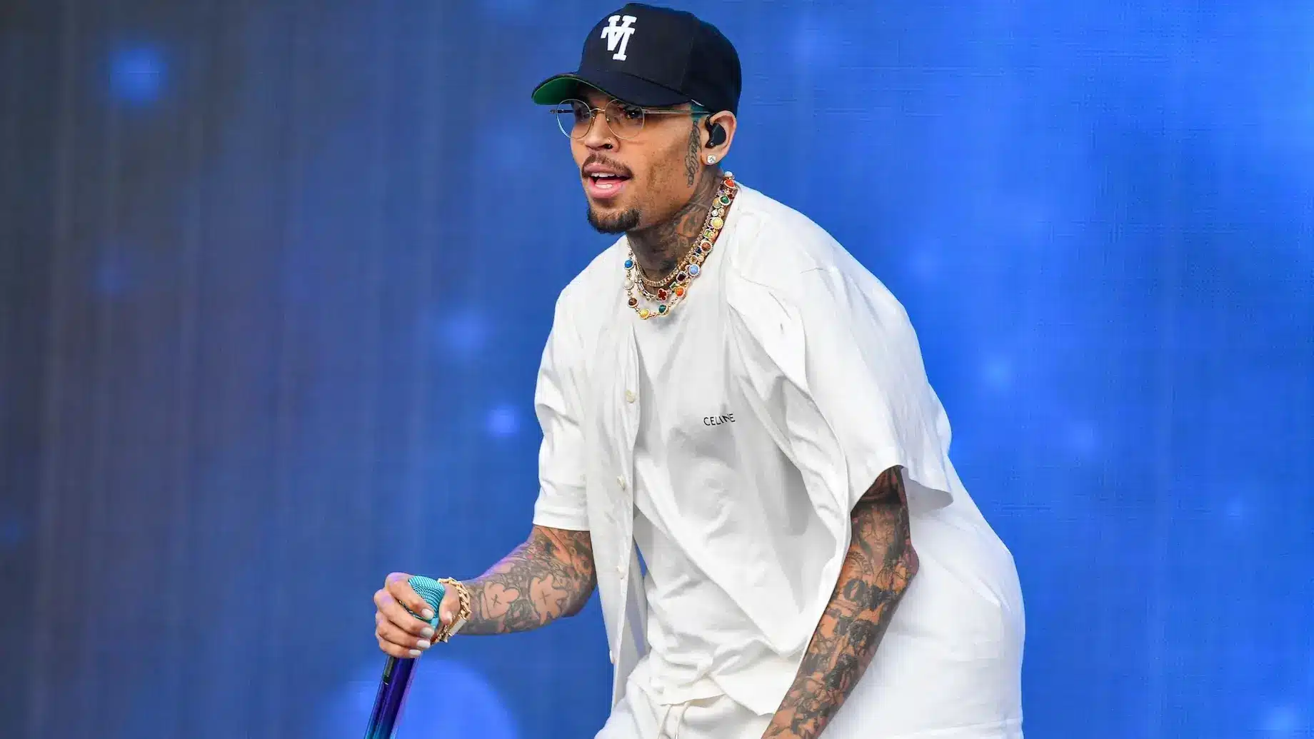 Chris Brown, A man with tattoos, wearing a navy blue baseball cap with "V" on it, glasses, a white short-sleeved shirt, and a white undershirt, performs on stage against a blue background, holding a purple microphone. He has multiple necklaces and bracelets.