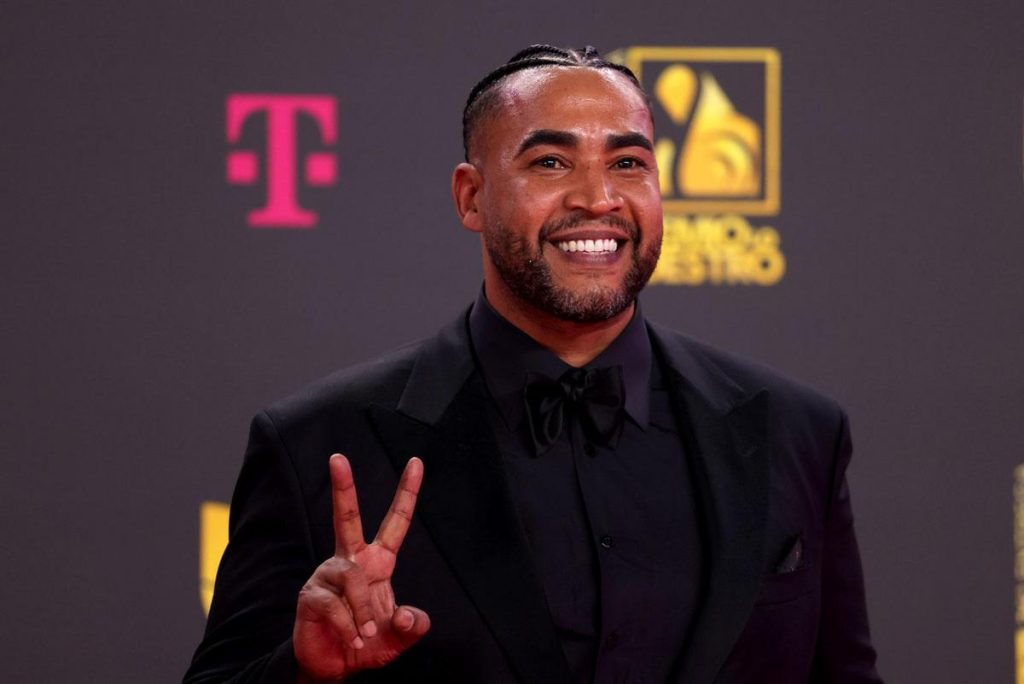 Don Omar, A man with a neatly trimmed beard and cornrows is smiling broadly on a red carpet. He is dressed in a black suit with a matching shirt and bow tie, holding up two fingers in a peace sign. Behind him, a dark background displays a T-Mobile logo in pink and a yellow and white event logo.