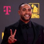 Don Omar, A man with a neatly trimmed beard and cornrows is smiling broadly on a red carpet. He is dressed in a black suit with a matching shirt and bow tie, holding up two fingers in a peace sign. Behind him, a dark background displays a T-Mobile logo in pink and a yellow and white event logo.