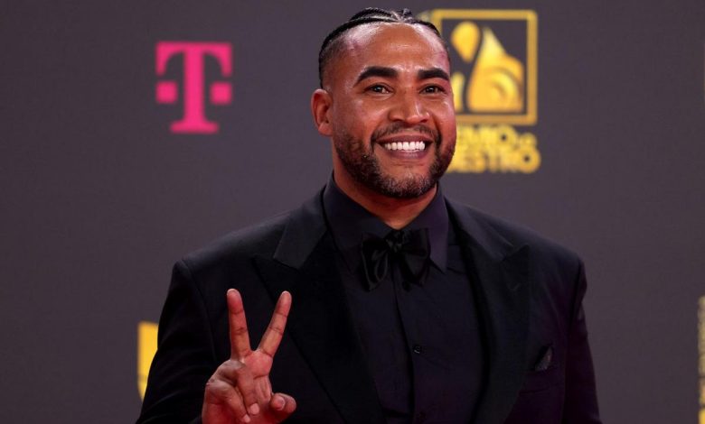 Don Omar, A man with a neatly trimmed beard and cornrows is smiling broadly on a red carpet. He is dressed in a black suit with a matching shirt and bow tie, holding up two fingers in a peace sign. Behind him, a dark background displays a T-Mobile logo in pink and a yellow and white event logo.