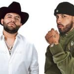 Two men pose against a white background. The man on the left wears a black cowboy hat, white shirt, and chain necklaces. The man on the right wears a black beanie, green hoodie, chain necklace, watch, and has visible tattoos on his hand and wrist.