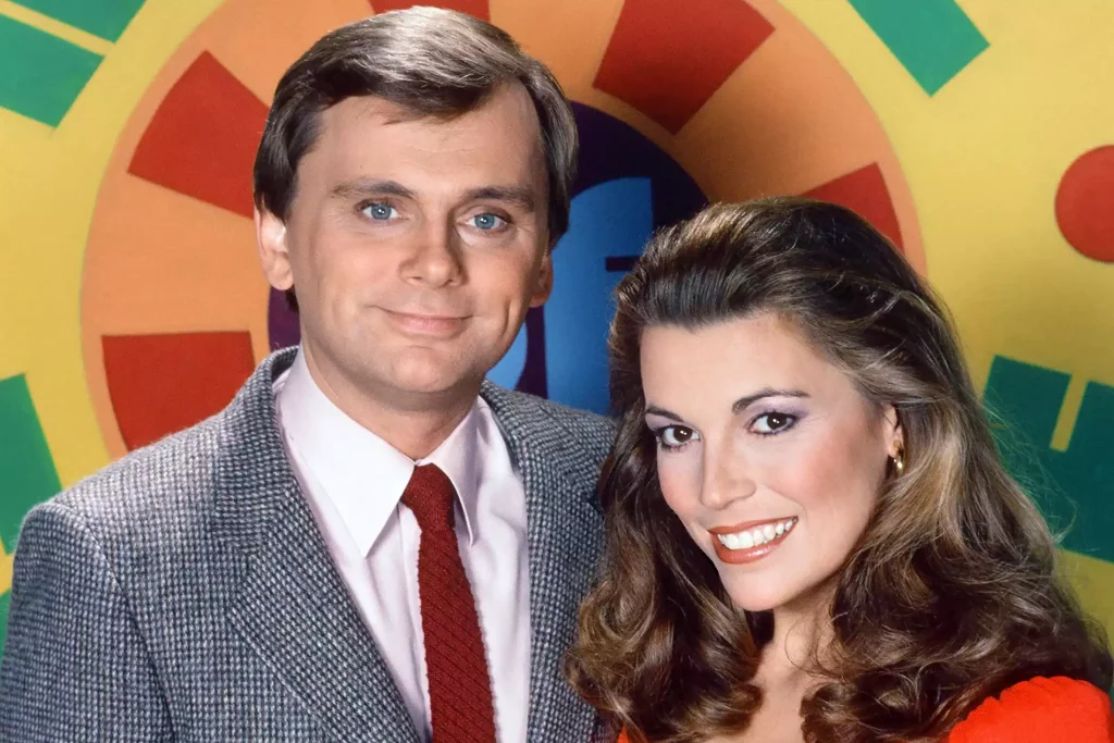 Pat Sajak (left) and Vanna White (right), A man and a woman are posing together in front of a colorful background featuring segments of green, red, and yellow. The man, with short brown hair, wears a gray suit, white shirt, and red tie. The woman, with long wavy brown hair, wears a bright red top and smiles warmly at the camera.