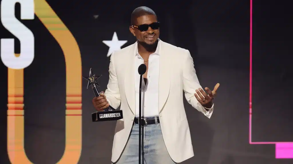 Usher, A man in a white blazer, light blue jeans, and black sunglasses stands on stage, holding a star-shaped award and speaking into a microphone. The background features a large illuminated design and a star symbol. He appears to be smiling and gesturing with his right hand.