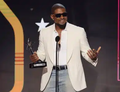 Usher, A man in a white blazer, light blue jeans, and black sunglasses stands on stage, holding a star-shaped award and speaking into a microphone. The background features a large illuminated design and a star symbol. He appears to be smiling and gesturing with his right hand.