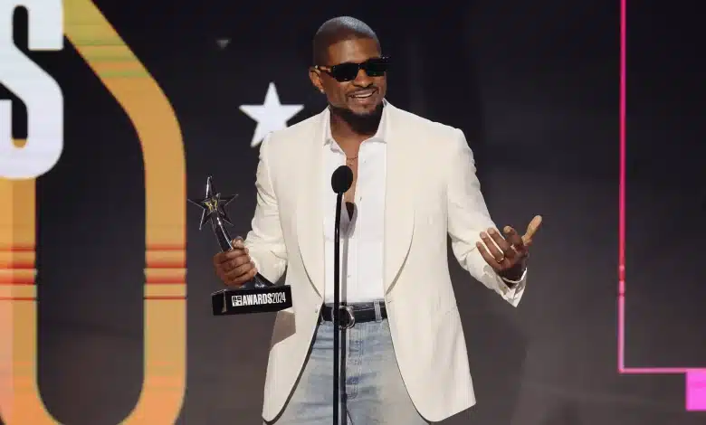 Usher, A man in a white blazer, light blue jeans, and black sunglasses stands on stage, holding a star-shaped award and speaking into a microphone. The background features a large illuminated design and a star symbol. He appears to be smiling and gesturing with his right hand.