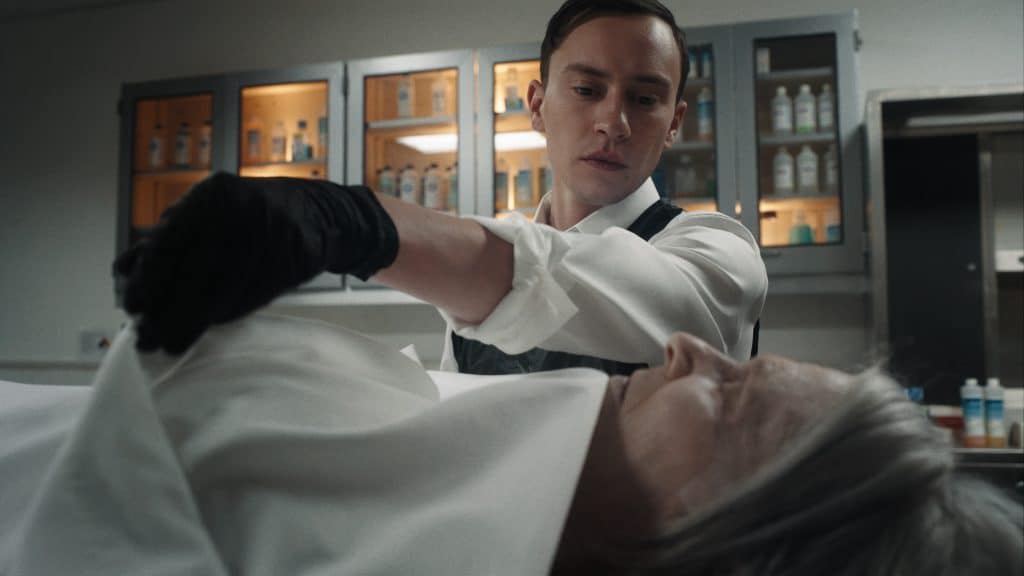 Kier Gilchrist and Annie Abbott, A man wearing a white shirt and black gloves examines the body of an older person draped in a white cloth in a clinical setting. Behind him, shelves with bottles and medical supplies are visible, illuminated by soft lighting. He appears focused and professional in his demeanor.