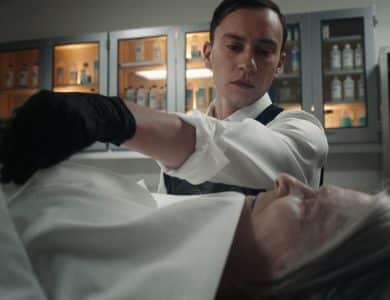 Kier Gilchrist and Annie Abbott, A man wearing a white shirt and black gloves examines the body of an older person draped in a white cloth in a clinical setting. Behind him, shelves with bottles and medical supplies are visible, illuminated by soft lighting. He appears focused and professional in his demeanor.