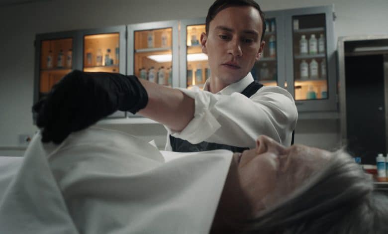 Kier Gilchrist and Annie Abbott, A man wearing a white shirt and black gloves examines the body of an older person draped in a white cloth in a clinical setting. Behind him, shelves with bottles and medical supplies are visible, illuminated by soft lighting. He appears focused and professional in his demeanor.