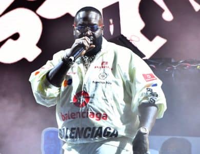 Rick Ross, A person wearing a white Balenciaga jacket, dark sunglasses, and multiple chains around their neck holds a microphone and performs onstage. Black and white abstract designs are projected on the background. The atmosphere is vibrant with dynamic lighting enhancing the performance.