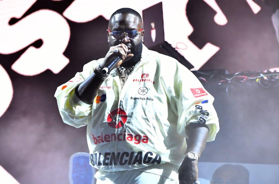 Rick Ross, A person wearing a white Balenciaga jacket, dark sunglasses, and multiple chains around their neck holds a microphone and performs onstage. Black and white abstract designs are projected on the background. The atmosphere is vibrant with dynamic lighting enhancing the performance.