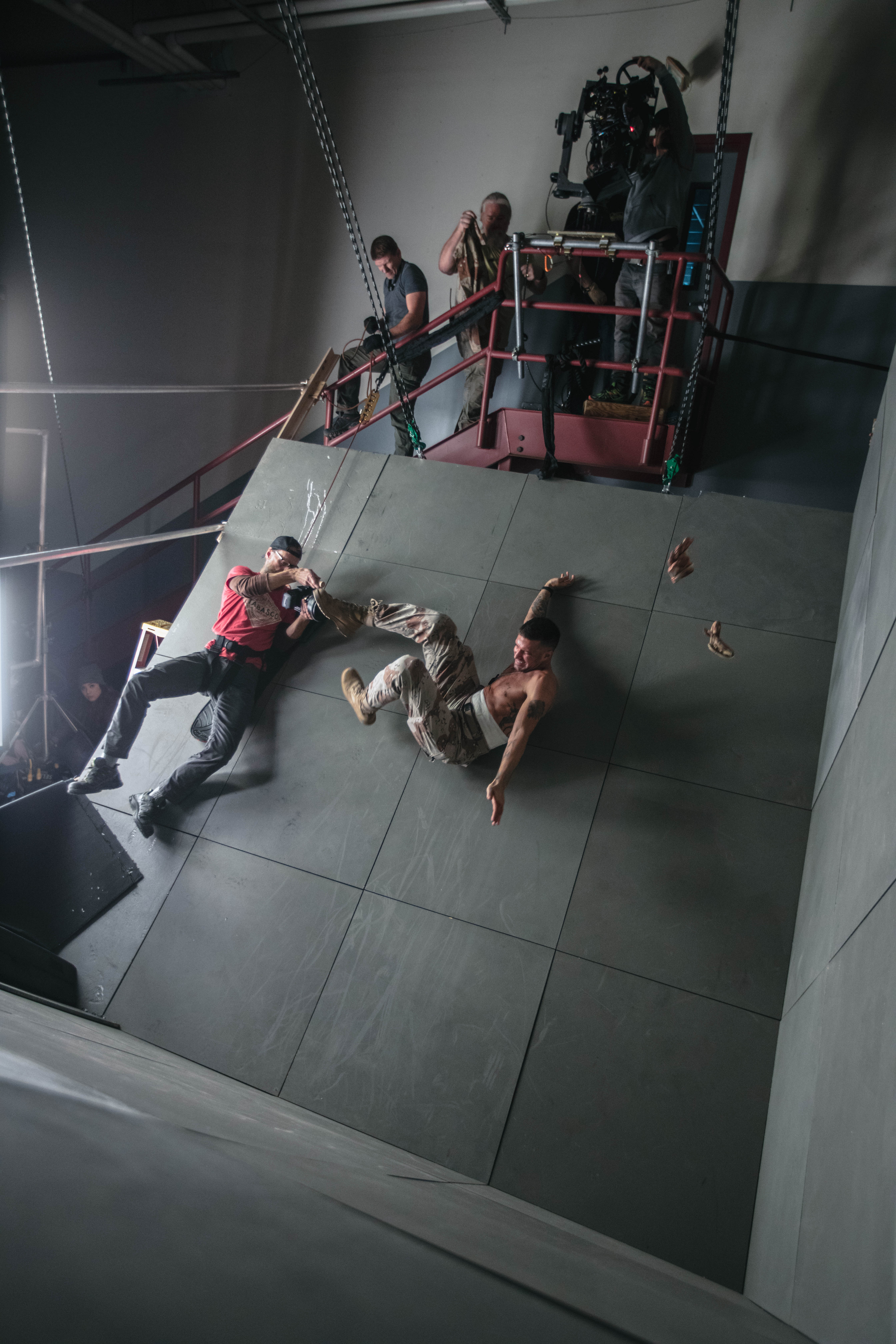 Two men in a staged action scene are suspended against a tilted wall. One man is kicking the other, who is falling backward. Both are equipped with safety harnesses. Crew members, including a cameraman on a raised platform, film the action from above. Various equipment is visible around the set.