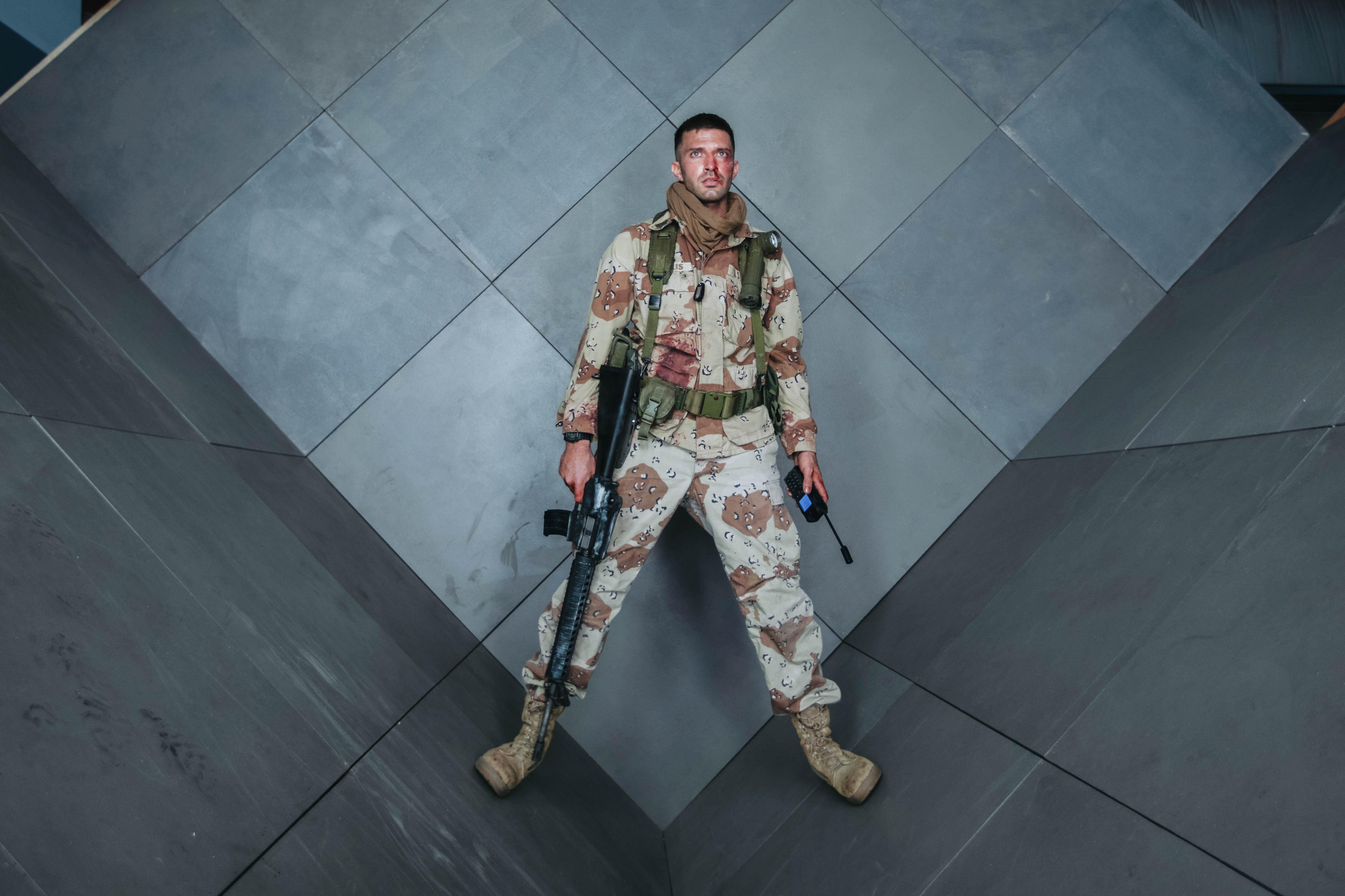 A soldier in camouflage stands with legs apart against a geometric, gray-tiled wall. He holds a rifle in one hand and a flashlight in the other. His face is smeared with dirt, and he has a serious expression. He wears tan combat boots, a helmet, and a tactical vest with various gear.