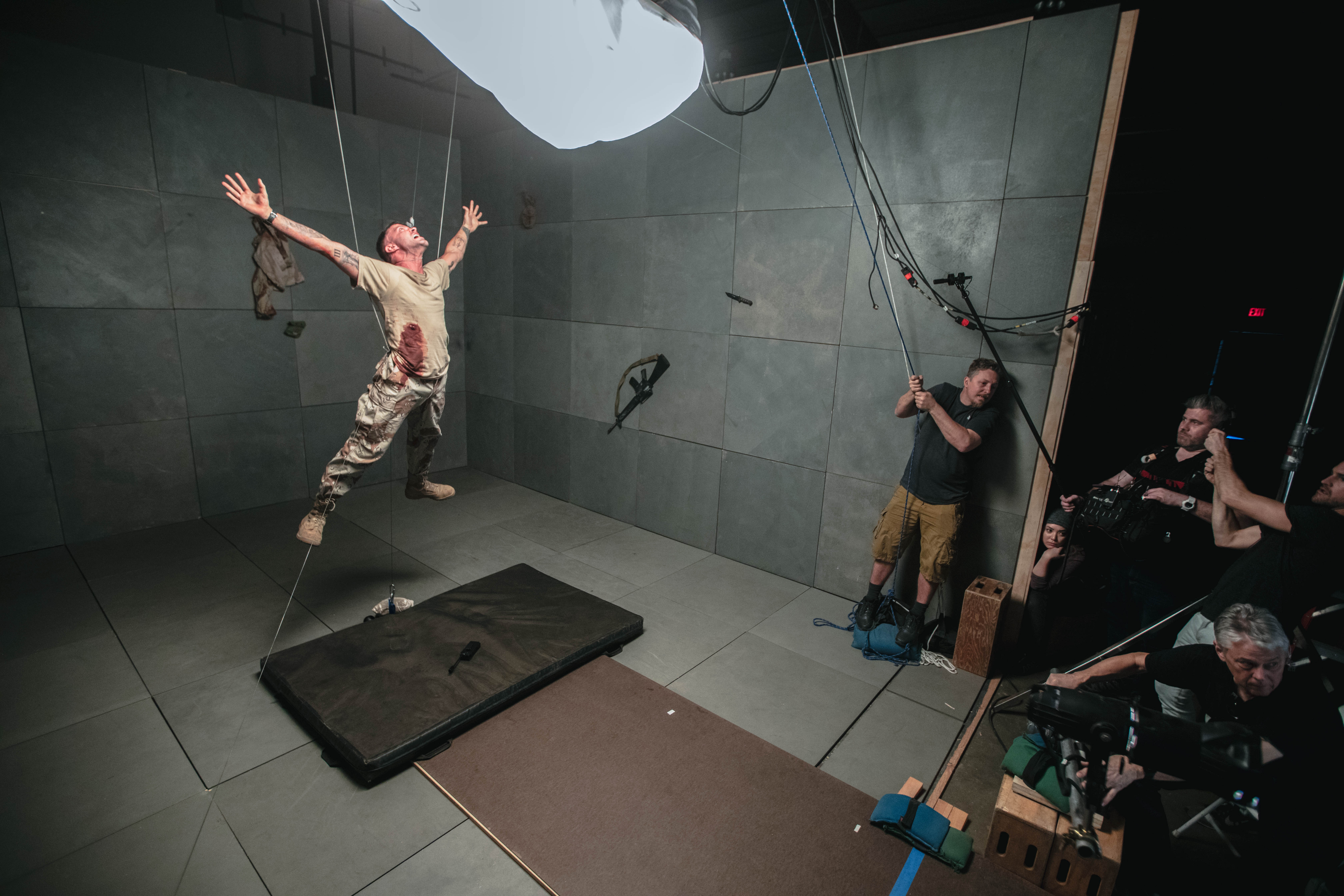 A person wearing a beige t-shirt and camouflage pants is suspended mid-air by wires in a studio setup. They are bleeding from the stomach with arms outstretched. Multiple crew members and cameras surround the scene, capturing the action. A thick mat lies on the floor beneath the suspended person.