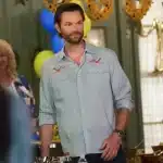 Jared Padalecki, A man with short brown hair and a beard, wearing a light blue shirt with embroidered patterns, stands indoors at a gathering. Behind him are festive decorations, including sunflower garlands and yellow and blue balloons. Two people in casual attire are visible in the background.