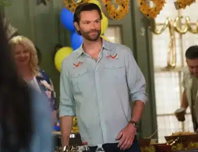 Jared Padalecki, A man with short brown hair and a beard, wearing a light blue shirt with embroidered patterns, stands indoors at a gathering. Behind him are festive decorations, including sunflower garlands and yellow and blue balloons. Two people in casual attire are visible in the background.