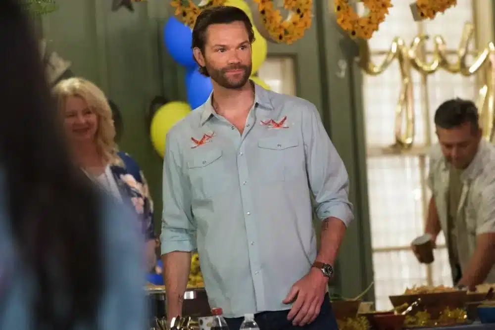 Jared Padalecki, A man with short brown hair and a beard, wearing a light blue shirt with embroidered patterns, stands indoors at a gathering. Behind him are festive decorations, including sunflower garlands and yellow and blue balloons. Two people in casual attire are visible in the background.