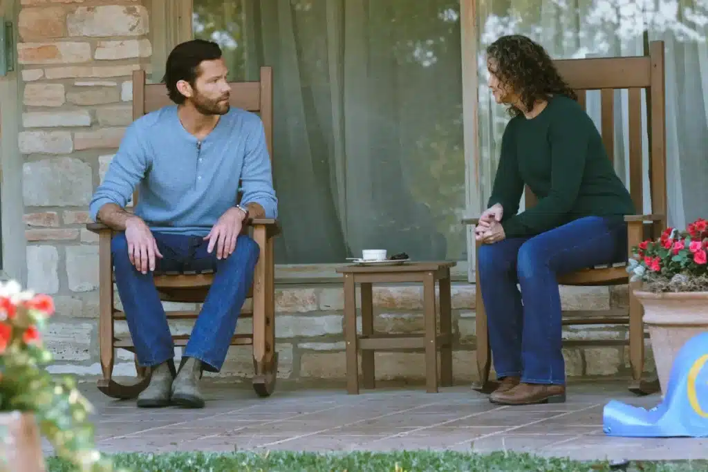 A man in a light blue long-sleeve shirt and jeans and a woman in a dark green sweater and jeans sit talking on wooden rocking chairs on a porch. Between them is a small table with a coffee cup, saucer, and smartphone. Flowers are in the foreground; beige stone wall and curtains are behind them.