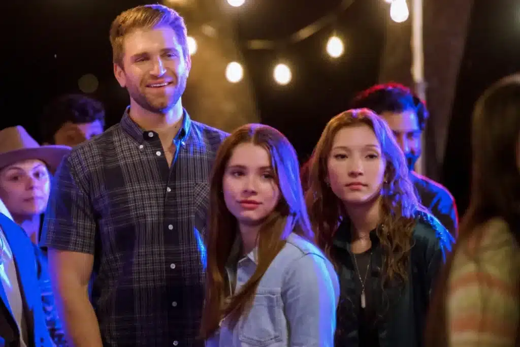 A young man with a beard and two young women with long hair, one in a light denim jacket and the other in a dark jacket, stand among a group of people outdoors at night, illuminated by string lights in the background. The scene has a vibrant, social atmosphere.