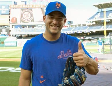 Jose "Candelita" Iglesias, A man at a baseball stadium, wearing a blue New York Mets cap and matching shirt, gives a thumbs-up with his right hand while holding a baseball glove. The background shows the field, other players, and a large screen displaying the Nationals Park logo.