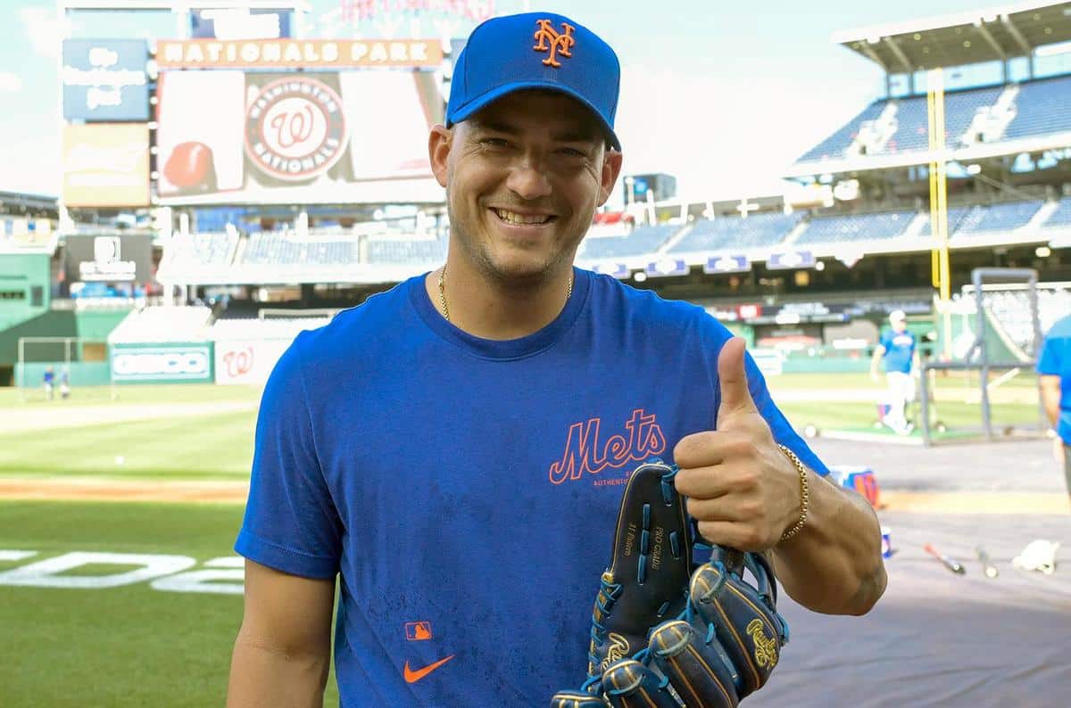 Jose "Candelita" Iglesias, A man at a baseball stadium, wearing a blue New York Mets cap and matching shirt, gives a thumbs-up with his right hand while holding a baseball glove. The background shows the field, other players, and a large screen displaying the Nationals Park logo.