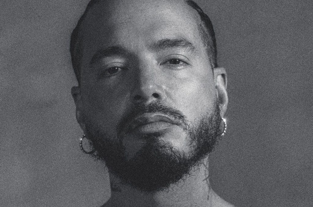 Black and white close-up portrait of a man with a trimmed beard and mustache, wearing hoop earrings. He has short, slicked-back hair and a neutral facial expression, looking directly at the camera. The background is plain and gray.