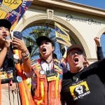 A dynamic group of people wearing SAG-AFTRA strike apparel stands passionately in front of the Paramount Pictures gate, holding signs and raising fists. A woman in a black shirt addresses the crowd through a microphone, flanked by two others in vests, all united in protest.