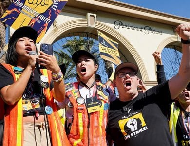 A dynamic group of people wearing SAG-AFTRA strike apparel stands passionately in front of the Paramount Pictures gate, holding signs and raising fists. A woman in a black shirt addresses the crowd through a microphone, flanked by two others in vests, all united in protest.