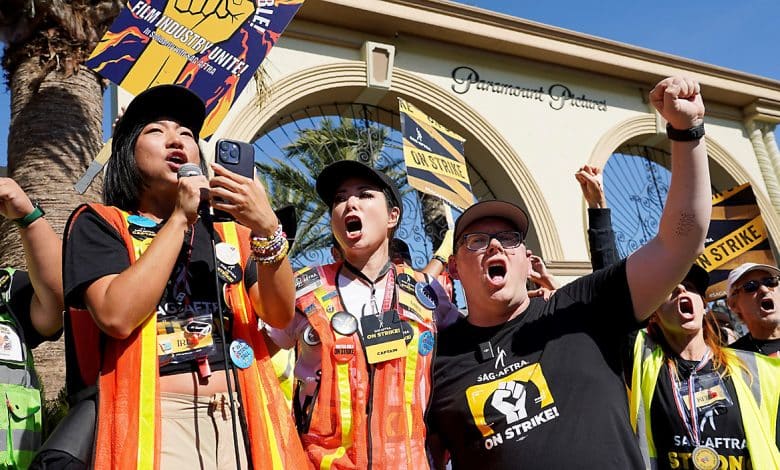 A dynamic group of people wearing SAG-AFTRA strike apparel stands passionately in front of the Paramount Pictures gate, holding signs and raising fists. A woman in a black shirt addresses the crowd through a microphone, flanked by two others in vests, all united in protest.