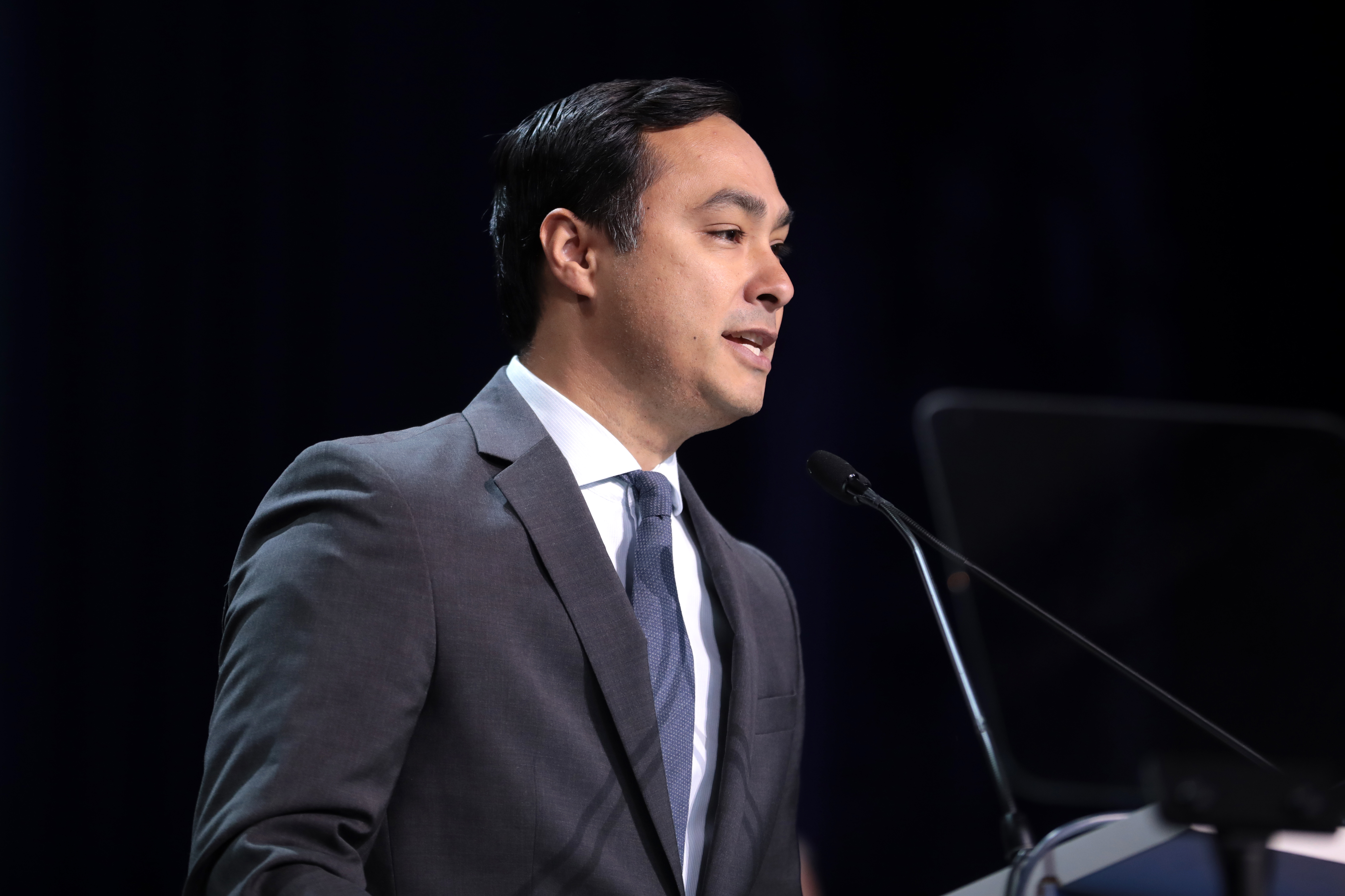 U.S. Congressman Joaquin Castro, A man in a gray suit, white shirt, and blue tie speaks into a microphone against a dark background. He has short, dark hair and is standing at a podium. The lighting highlights his face and upper body as he passionately addresses the audience about Latino music nominations and their cultural impact.