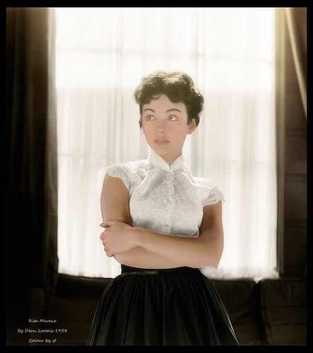 Rita Moreno, A woman in a vintage style stands with arms crossed, wearing a short-sleeve white lace high-neck blouse and a high-waisted black skirt. She has short dark hair and a contemplative expression. The background features a softly lit window with sheer curtains, creating a nostalgic ambiance.