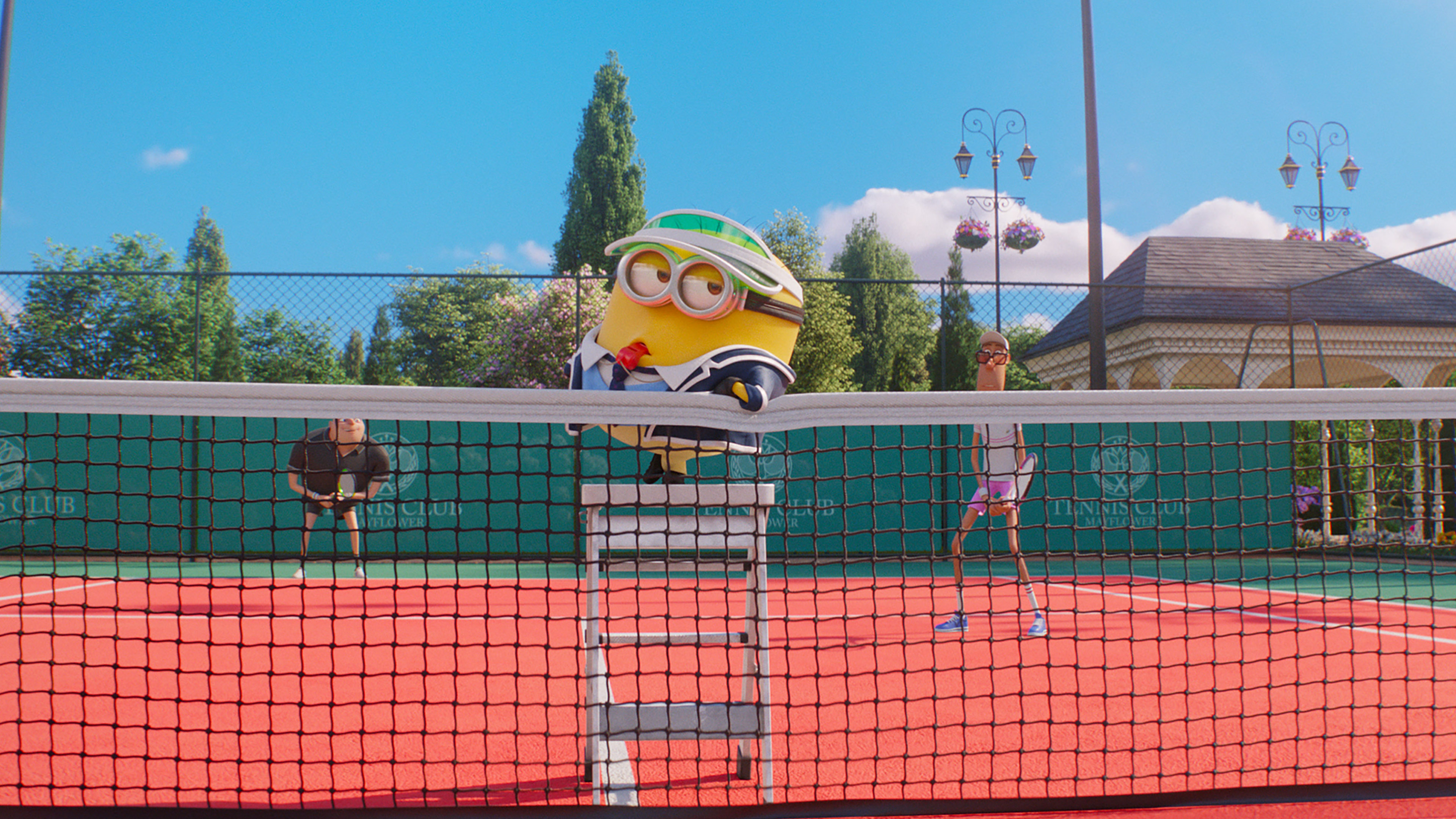 A scene from a Minions movie featuring a Minion dressed as a tennis umpire, sitting on a chair at the tennis net. Two players are on a red tennis court ready to play, with trees, a clubhouse, and a bright blue sky in the background. The Minion wears a green visor and sunglasses, adding to the hilarious antics typical of Universal Pictures' Minion Mini-Movies.