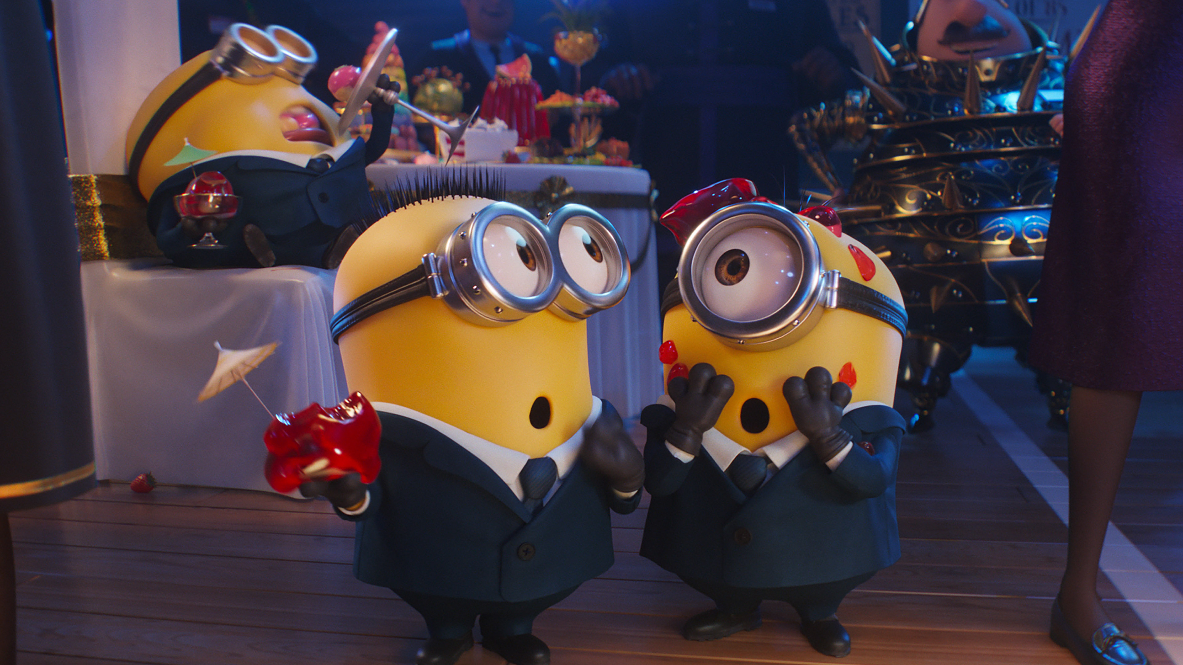 Two Minions in suits are depicted at a festive party. One Minion holds red fruits, surprised, while another has food on its goggles and head. In the background, a third Minion sprawls on a table covered with food, looking delighted. This scene feels straight out of one of those hilarious Minion Mini-Movies by Universal Pictures—a perfect family movie moment!