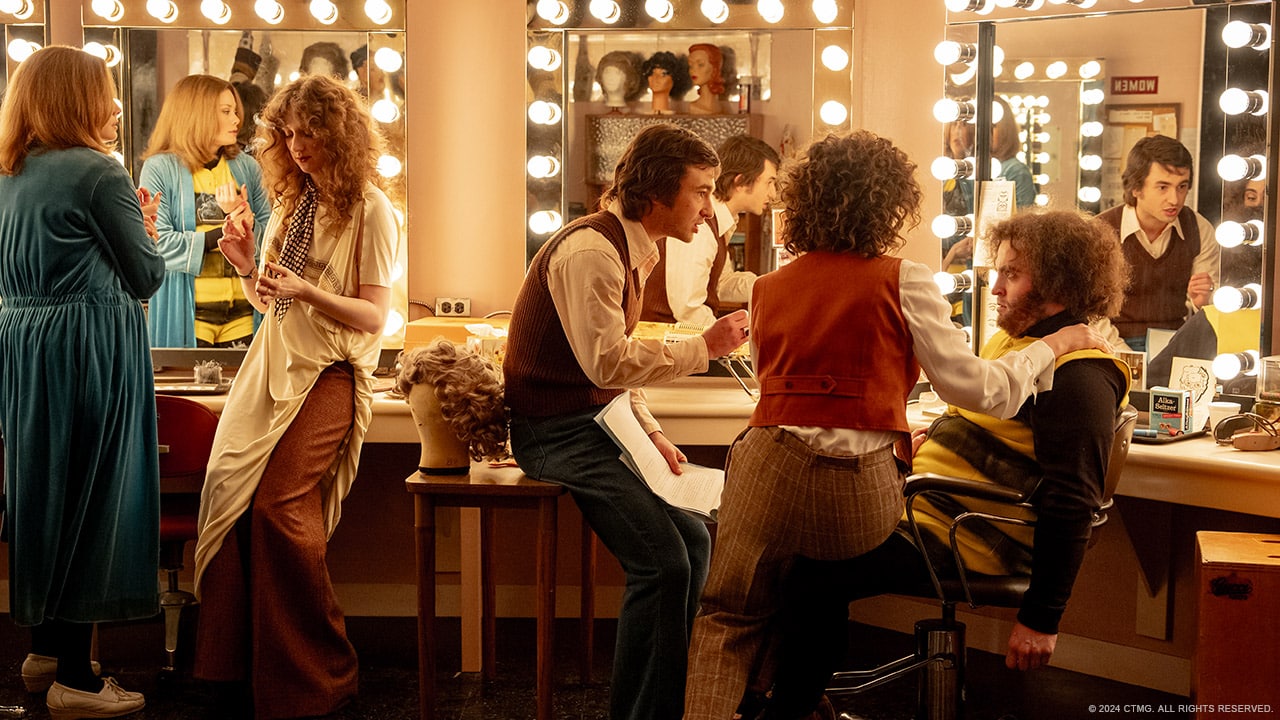 A brightly lit backstage dressing room filled with people. Four individuals are getting styled in front of makeup stations with illuminated mirrors. Two stylists are attending to clients, while others chat or prepare. Wigs and various styling tools are visible on the counters.