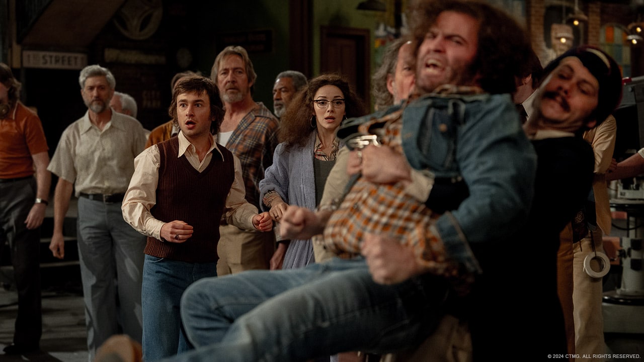 A group of people dressed in retro 1970s clothing are indoors, looking surprised and concerned. A woman in glasses and a man in a sweater vest are at the forefront of the group. Two men are seen lifting another man who appears unconscious or incapacitated, creating a scene of distress.