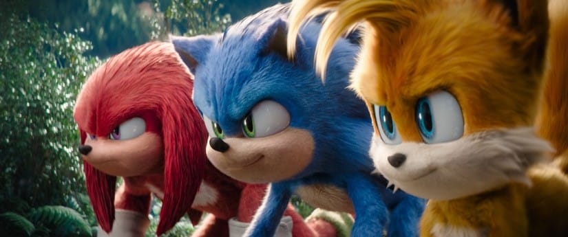 Three animated characters from the Sonic the Hedgehog series stand ready for action. From left to right: Knuckles, with red fur and white gloves; Sonic, with blue fur and white gloves; and Tails, with yellow fur, white gloves, and two tails. They are in a lush, green, forest-like environment.