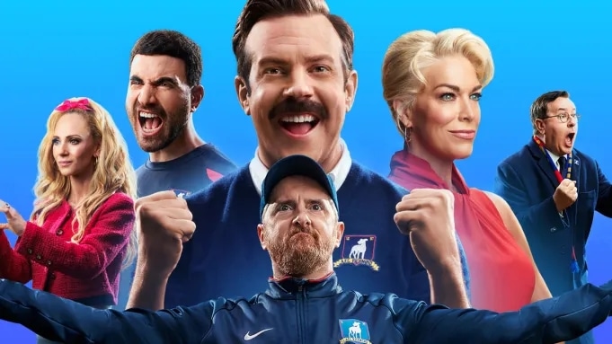 A colorful promotional image featuring six characters from the TV show, Ted Lasso, set against a blue background. Front and center is a mustachioed man in a blue sports jacket, flanked by cheering and smiling individuals, including two women and three men, suggesting team spirit and enthusiasm.