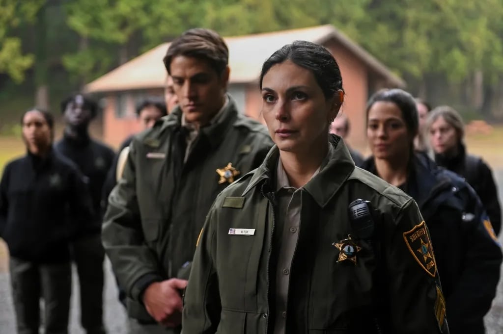 Morena Baccarin and a group of uniformed law enforcement officers stands outdoors with a cabin and trees in the background. A woman in the foreground, dressed in a green sheriff's jacket with a badge, looks serious. Other officers, both male and female, stand behind her in focus and blurred.