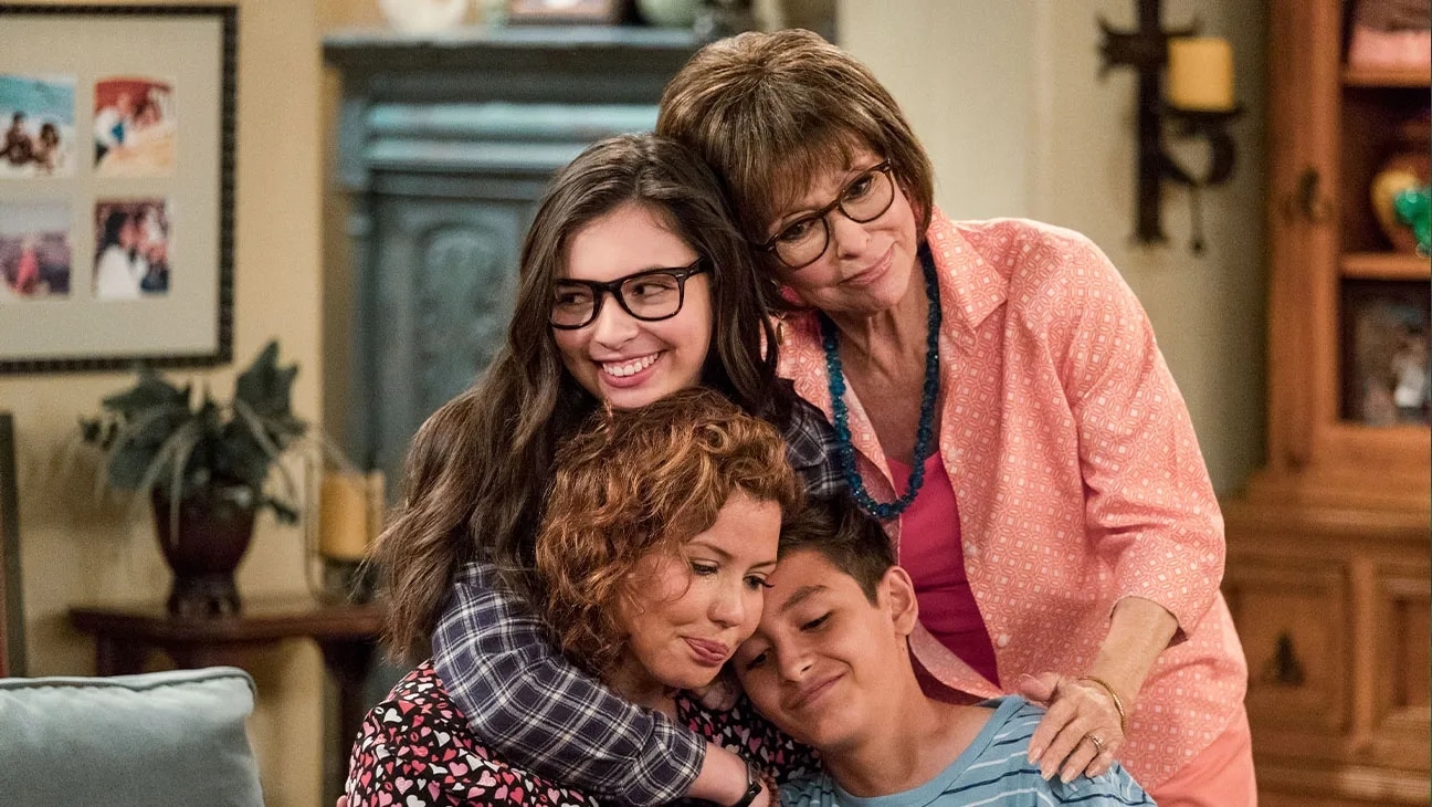 The cast of Netflix's "One Day at a Time", A group of four people shares a warm embrace in a cozy, well-decorated living room. An elderly woman with glasses stands behind, smiling. A young girl with glasses and wavy hair also smiles while hugging two others: a curly-haired woman and a boy in a striped shirt, who both look content.